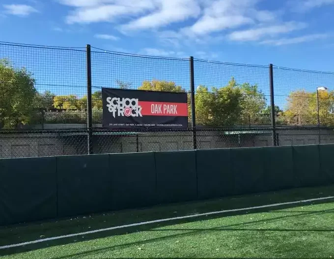 A banner for School of Rock Oak Park hangs on a fence overlooking a green turf field.