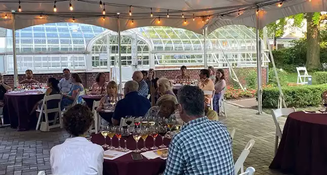 Guests seated under a tent at tables with wine glasses, with a greenhouse in the background.