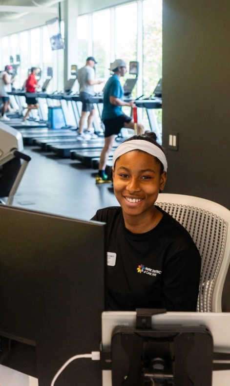 Smiling young woman at a computer with a gym and treadmills in the background.