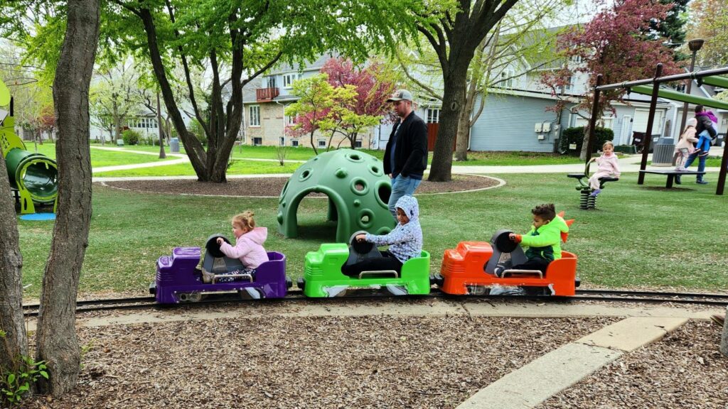 three children hand pedal a purple, green, and red train car