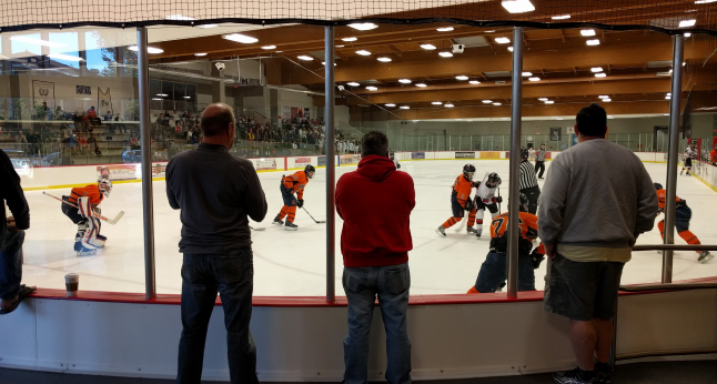 Spectators watch a youth hockey game from behind glass. Players in orange and white uniforms compete on the ice.
