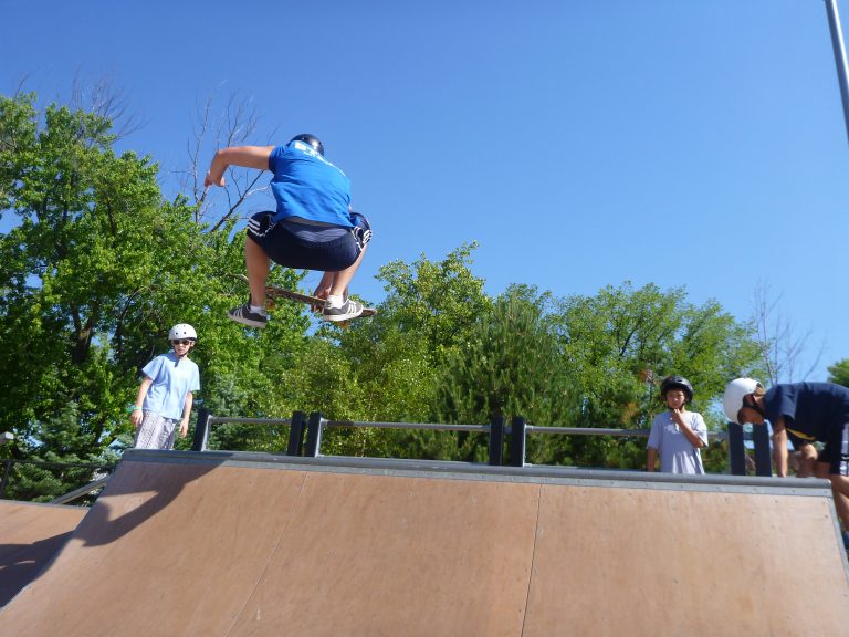 Teenager in blue shirt and shorts performs a skateboard jump at a skate park, with other kids watching.