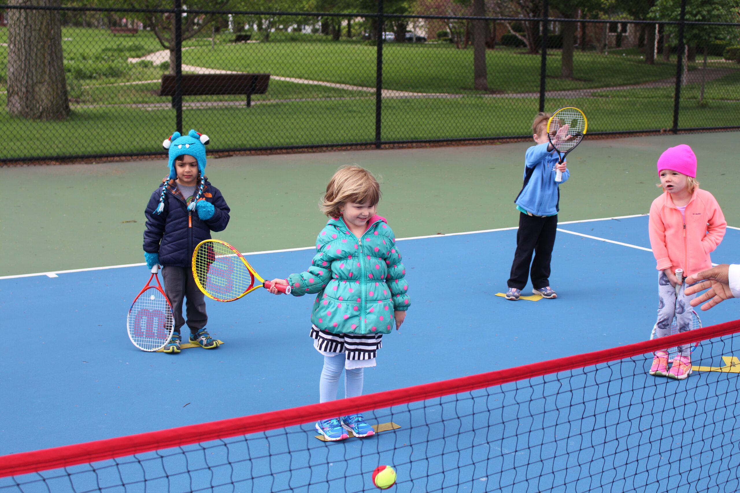 Four children on a blue tennis court, each holding a tennis racket. One child in the foreground is smiling and wearing a