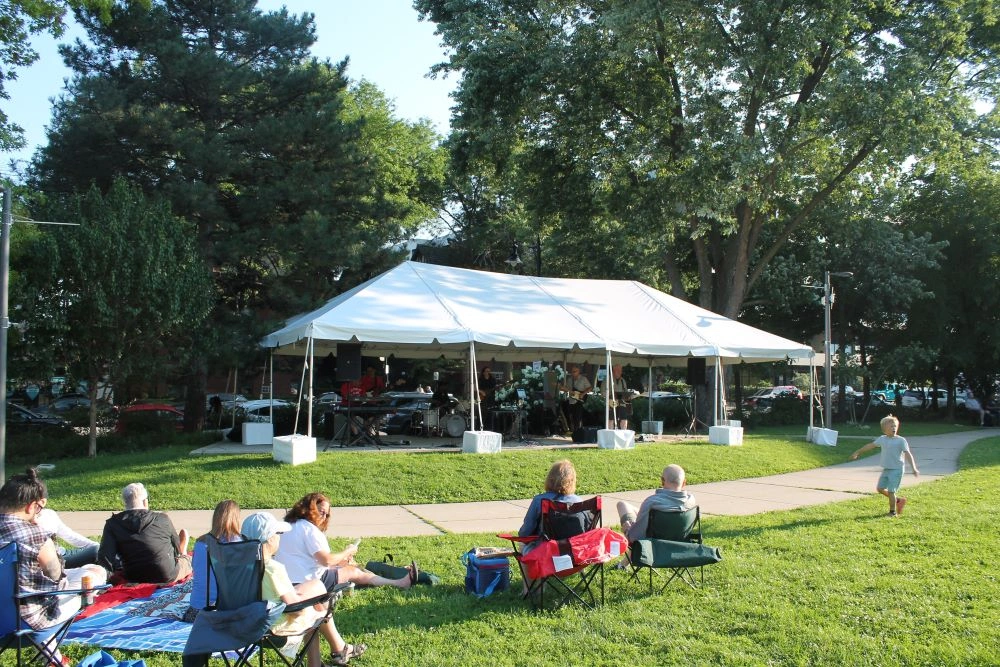 A band performs under a white tent at a park while an audience sits on the grass. A child runs on a path.