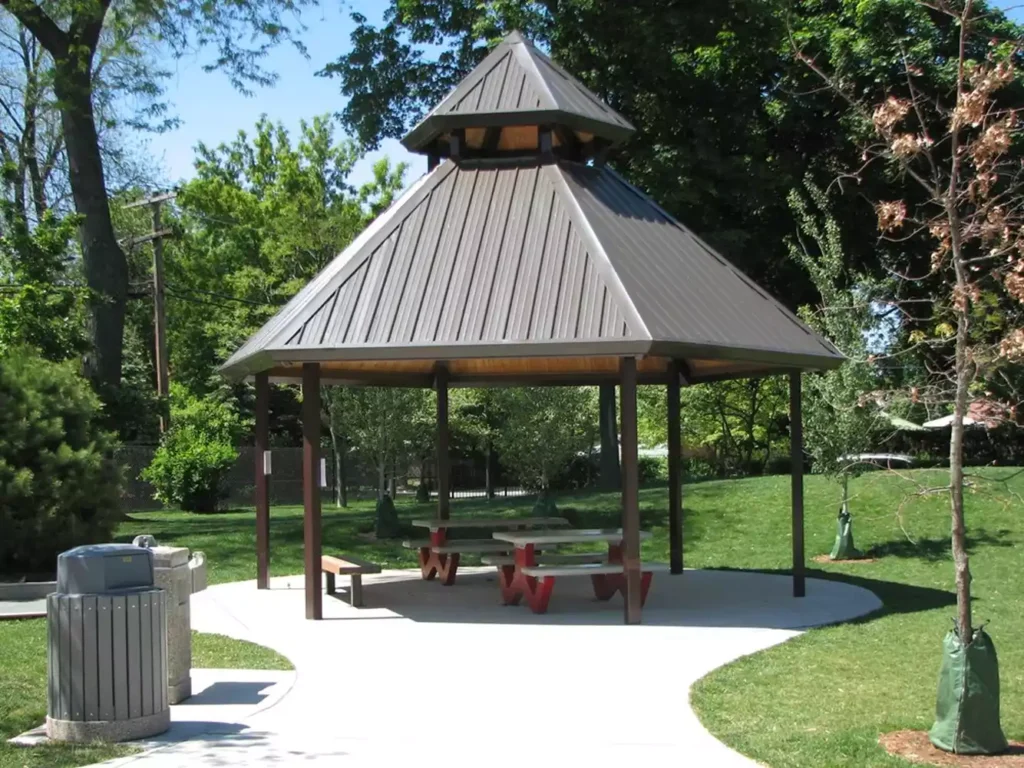 A metal-roofed gazebo with picnic tables and benches sits on a paved area in a park, surrounded by grass and trees.
