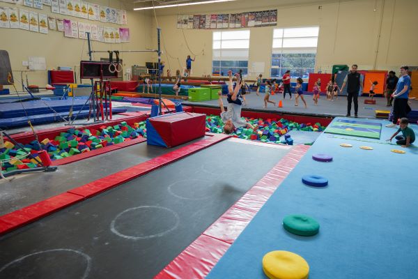 a boy does a flip on a tumble track with gymnasts taking class on the floor behind