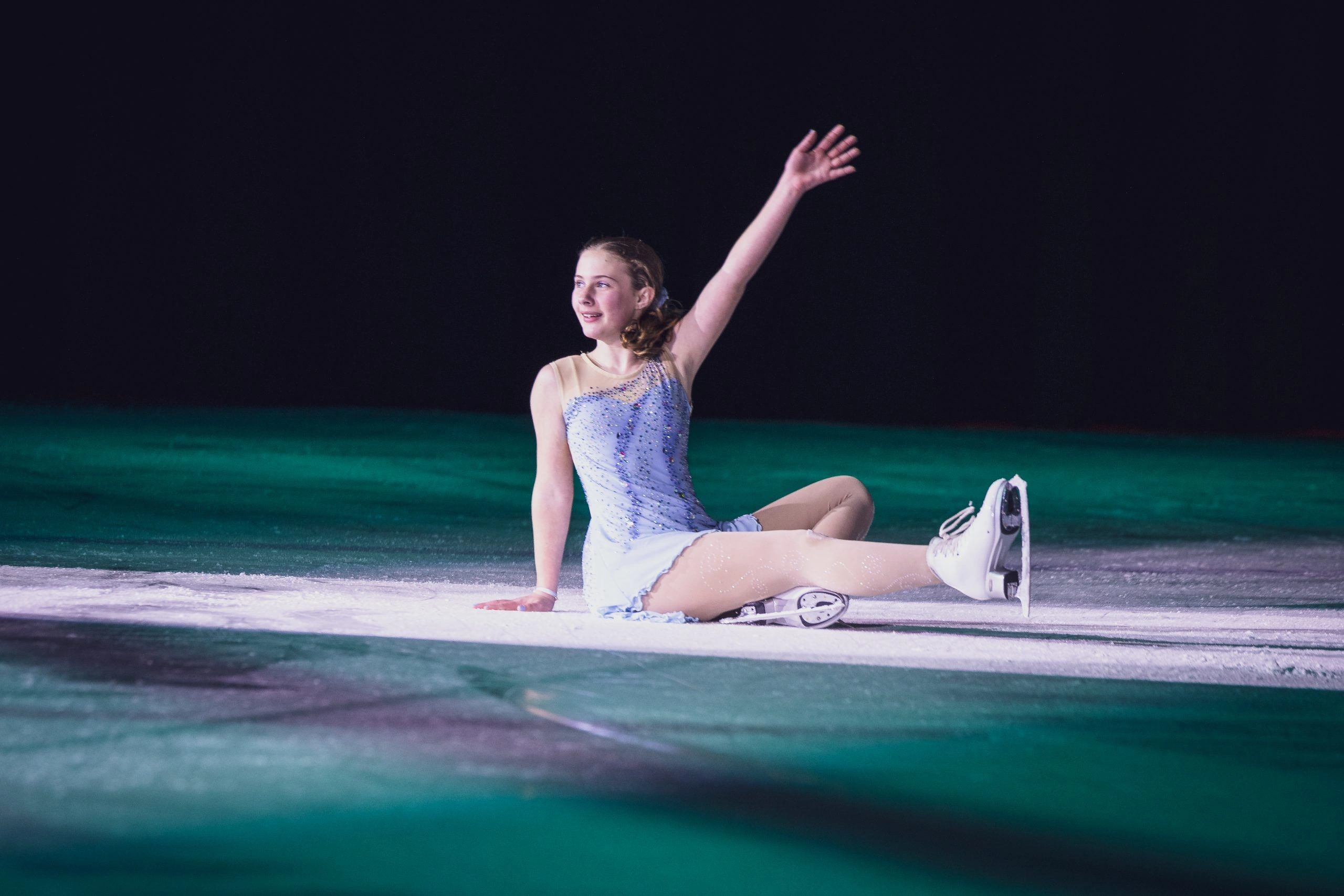 A young figure skater in a sparkly blue costume sits on the ice with one leg extended and her arm raised.