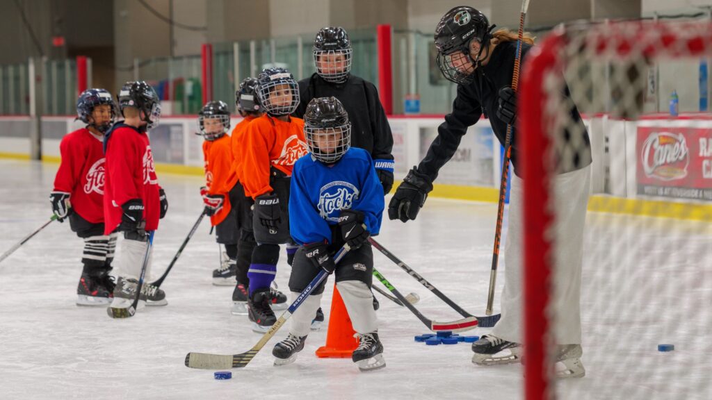 a line of young hockey players face the net while the coach points out their stance