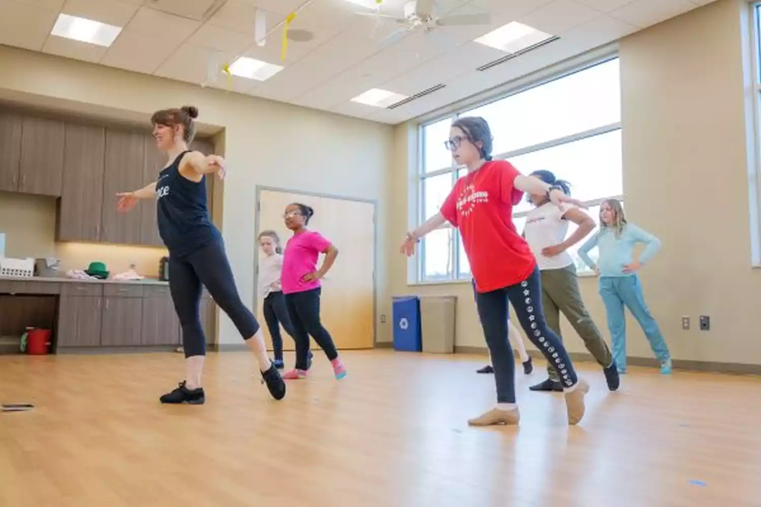 A dance instructor leads a group of young students in a ballet class, demonstrating a pose on a wooden floor in a studio.
