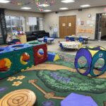 wide view of the cubhouse indoor playground with a nature themed rug, toddler tables, climbing equipment, and toys